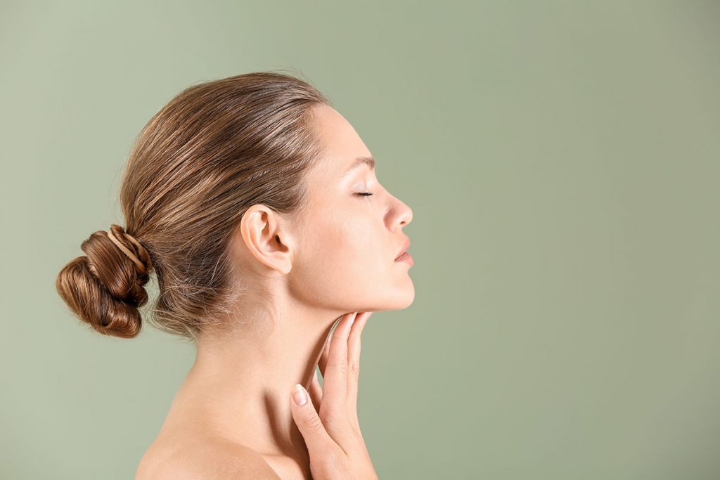 Woman with brown hair in a bun touches her neck, eyes closed, against a light green plastic surgery studio backdrop.