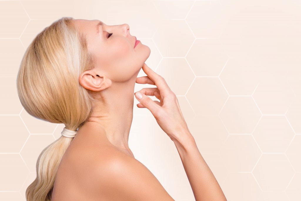 Woman with blonde hair in a ponytail tilts her head back, highlighting her chin against a beige hexagon-patterned background, evoking plastic surgery.