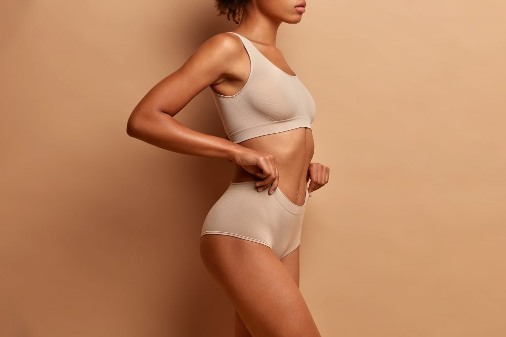 Woman in beige underwear set stands against a matching beige background, showcasing natural beauty post-plastic surgery.