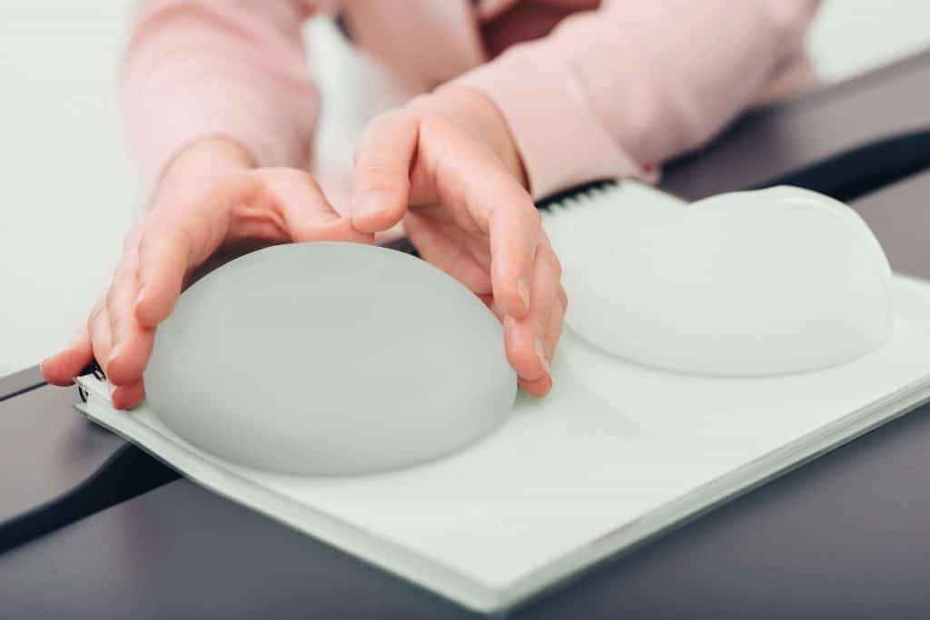 A person holds a silicone breast implant on a desk next to another implant. Both implants are sitting on a notebook.