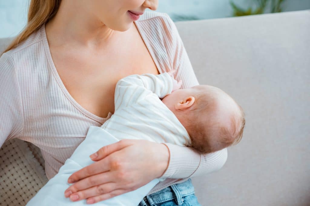 A woman sits on a couch cradling and breastfeeding an infant.