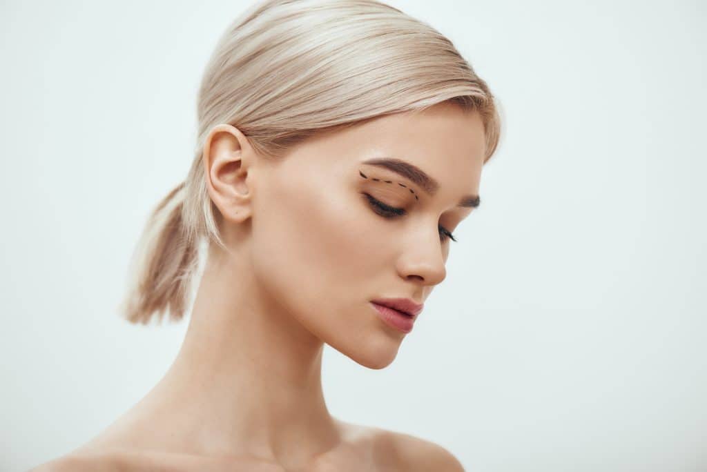 Woman with blonde hair in a low ponytail looking down, showing eyeliner and dotted plastic surgery lines near her eyelid.