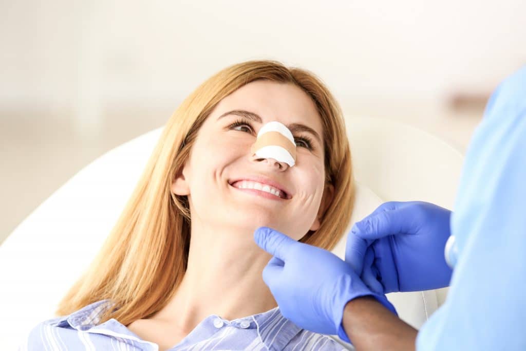 Smiling woman with nasal bandage is looking up at a person with gloved hands.