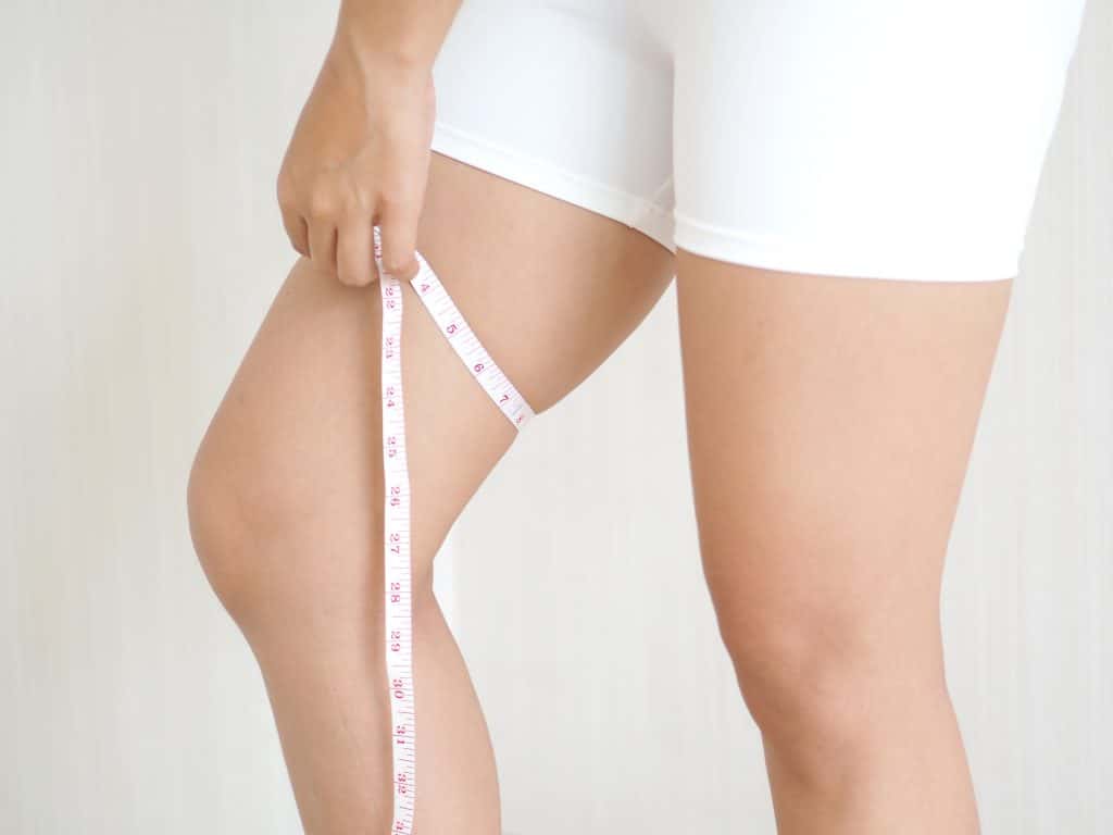 A person wearing white shorts measures the circumference of their upper thigh using a measuring tape.