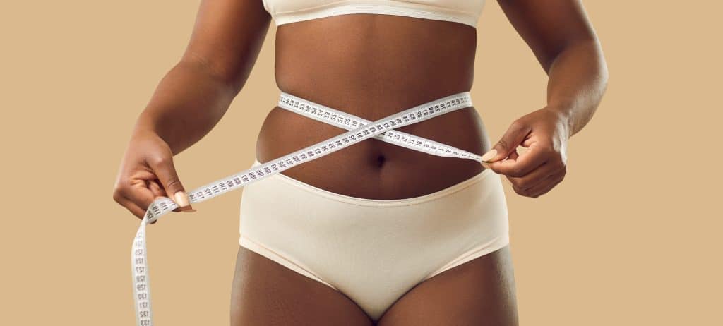 Woman in cream-colored undergarments measures her waist with a tape measure, set against a warm beige background.