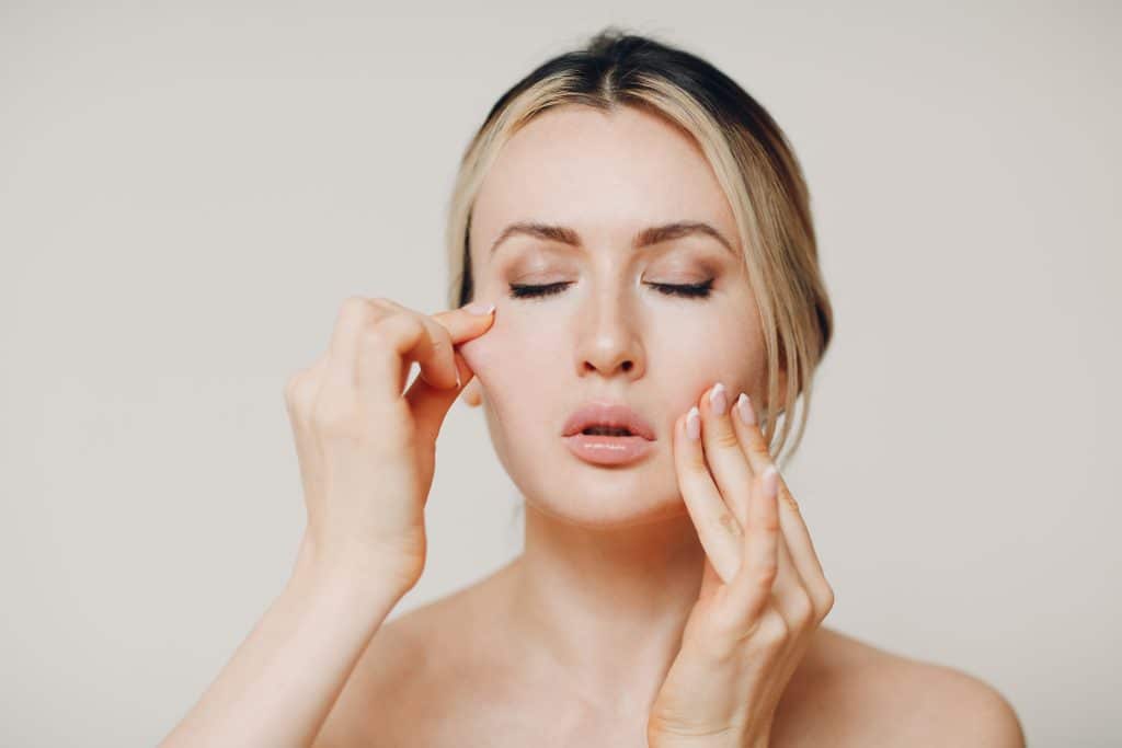 A close-up of a woman with her eyes closed, softly pulling the skin on her cheek with her fingertips.