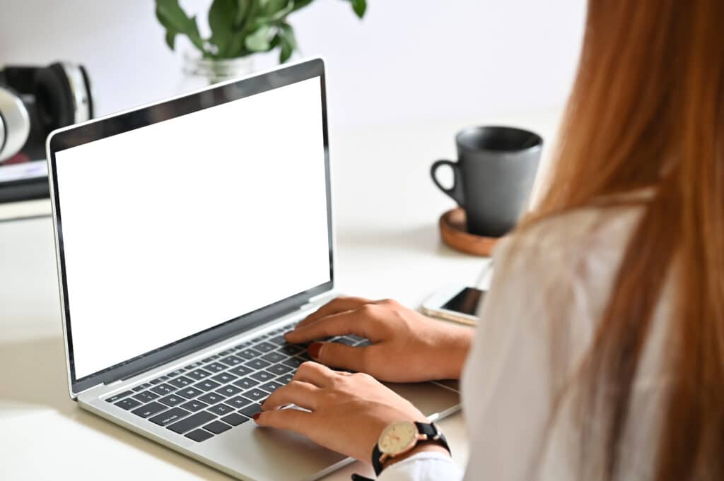 A person types on a laptop with a blank white screen at a desk.