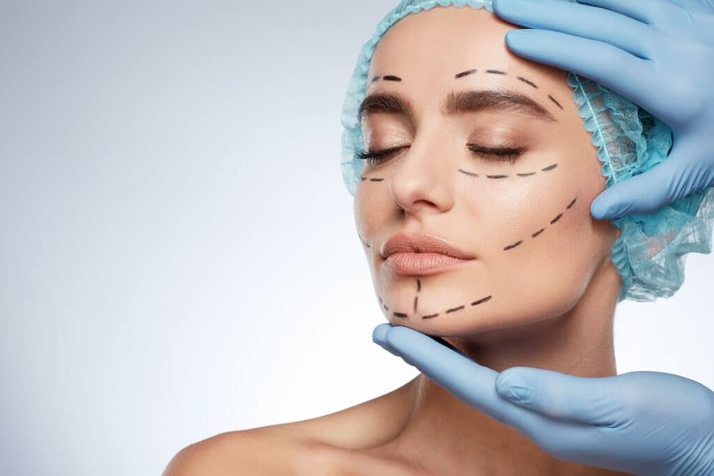 Woman wearing a surgical cap with dotted lines on her face, as gloved hands touch her face.