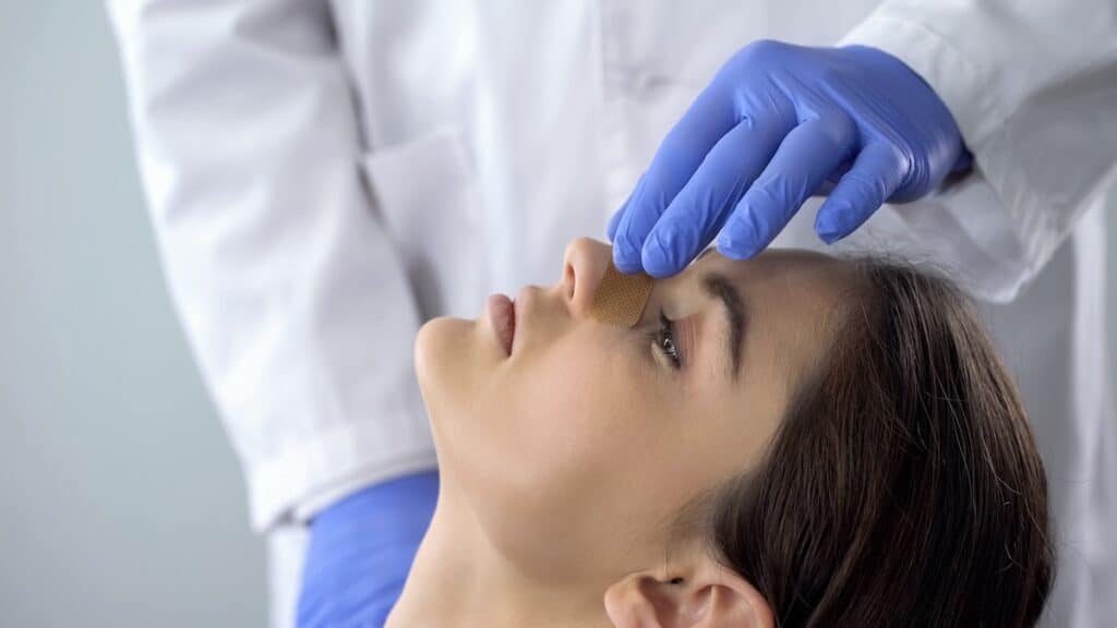 Surgeon in gloves applying a bandage to a patient’s nose during post-operative care.