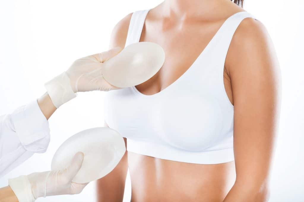 Gloved hands holding breast implants near a woman in a white sports bra.