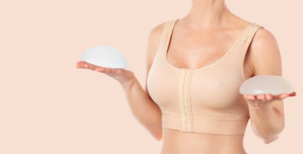 Woman wearing beige compressing bra after breast augmentation. Holding round implants.