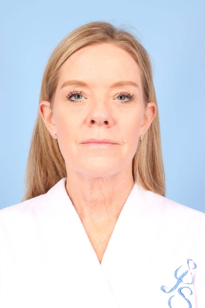 A woman with straight blonde hair wearing a white robe stands against a plain light blue background, facing forward.