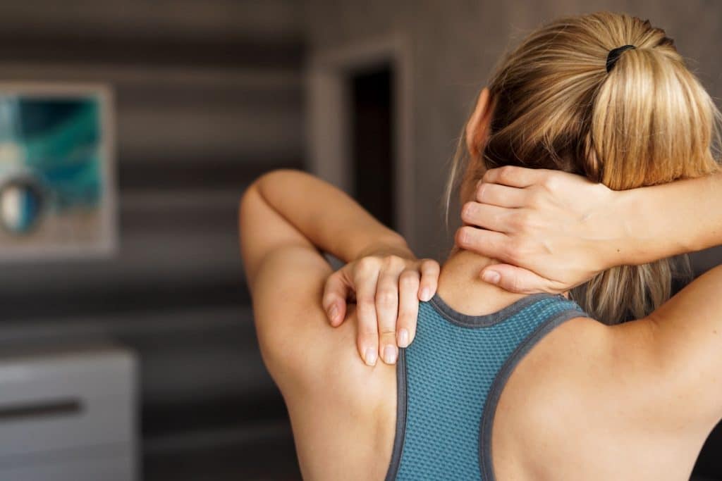 Woman in athletic wear seen from behind, holding her neck and upper back, appearing to have pain.