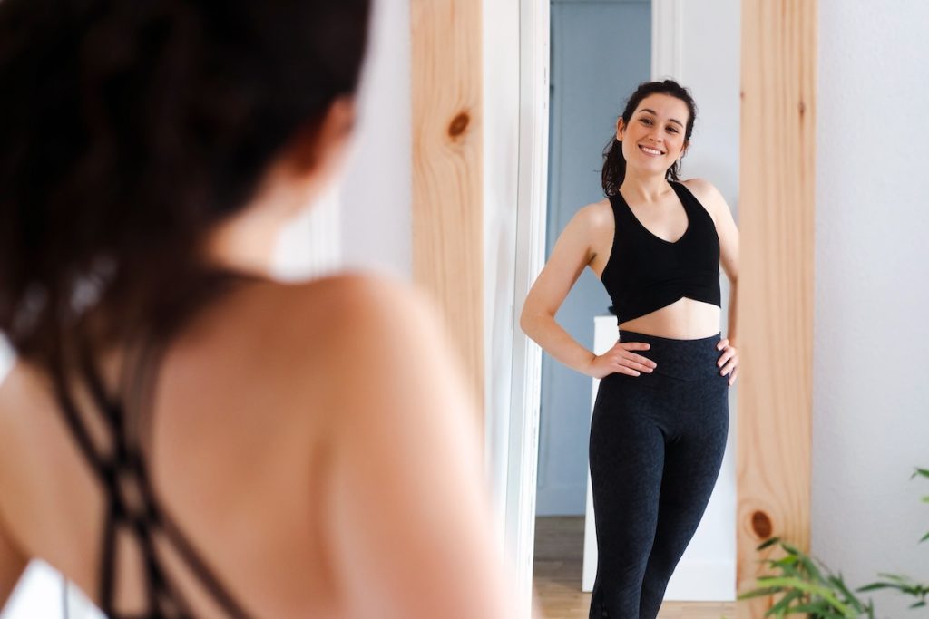 Woman in black workout clothes stands in front of a mirror, smiling with hands on hips in a bright room.