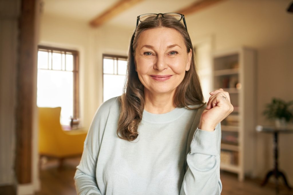Smiling middle-aged woman with glasses on her head sits indoors in a well-lit living room.