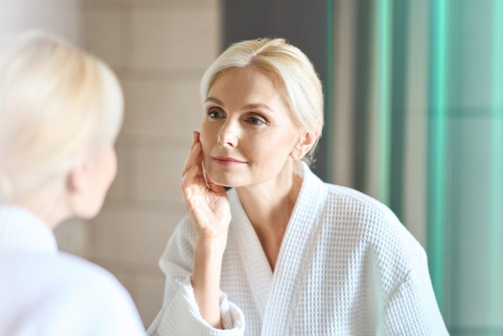 A woman in a white robe looks at her reflection in a mirror, touching her face with one hand.