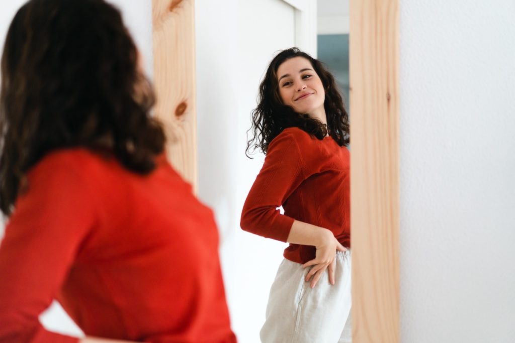 A woman in a red top and white pants smiles while looking at herself in a mirror.