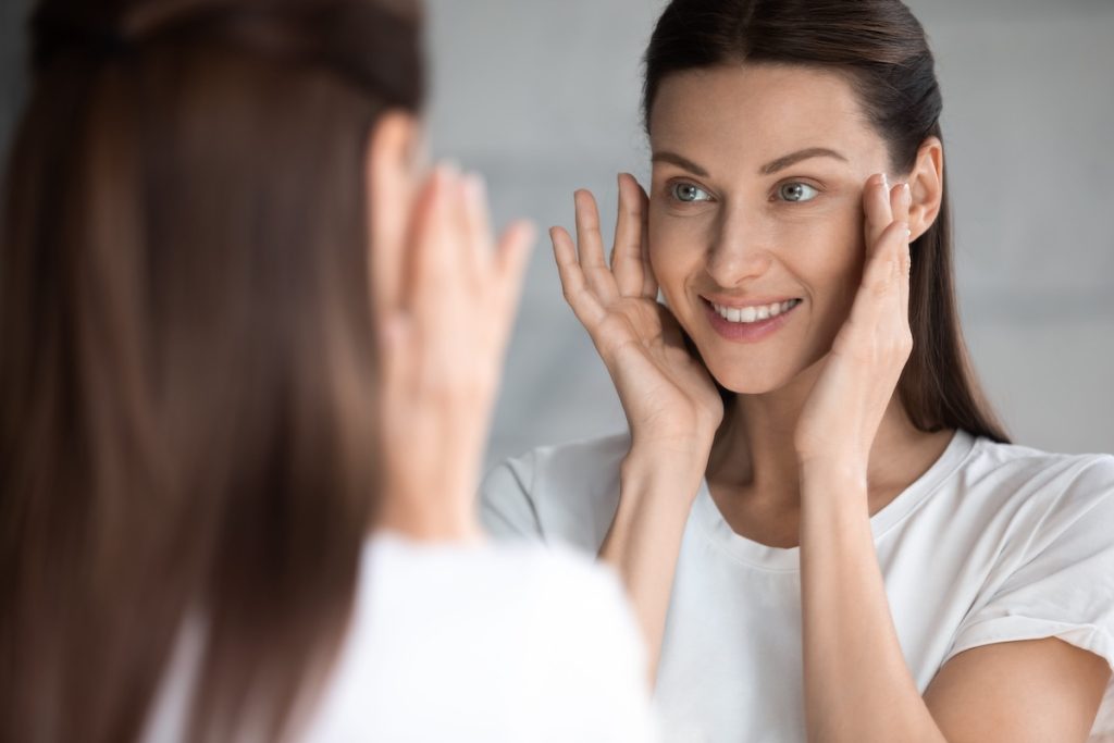 Woman in a white shirt smiles while gently touching her face and looking at herself in a mirror.