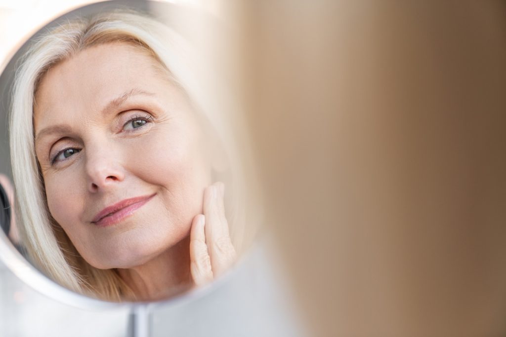 An older woman with blonde hair looks at her reflection in a mirror, gently touching her face with her hand.