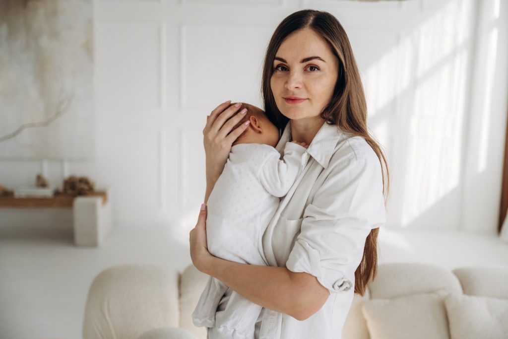 Woman in white shirt holding a baby against her chest in a bright, softly lit living room.