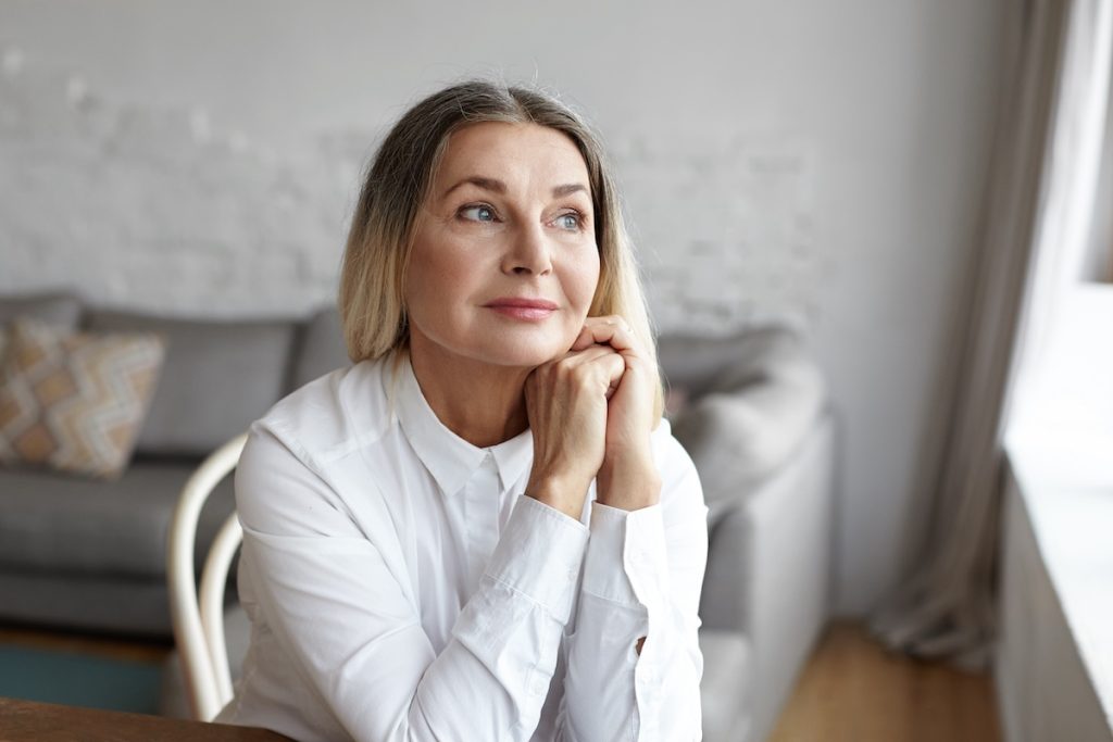 Woman with gray hair in a white shirt sitting indoors, looking thoughtfully out a window, hands resting under chin.